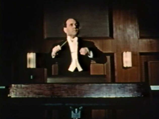 Frame from the 1956 educational film The Symphony Orchestra: a conductor in white tie raises his baton before a wood-paneled wall.