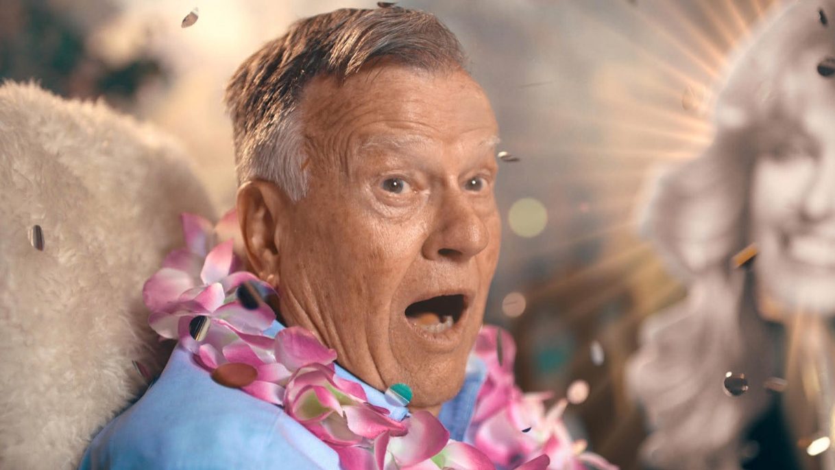Still from Dick Johnson Is Dead. Dick Johnson wears a flower lei, backlit and wide-eyed, in one of the film's staged afterlife sequences.