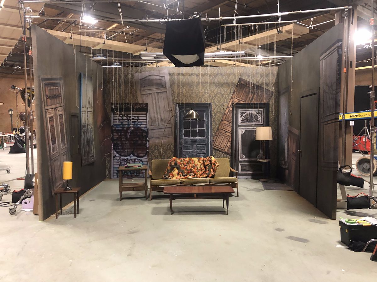 The hell set mid-build on a soundstage: walls of salvaged doors around a couch, a lamp and a coffee table.