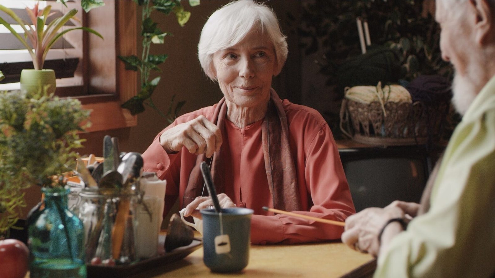 Still from Hello, World. An older woman sits at a diner counter with a mug, facing a companion.