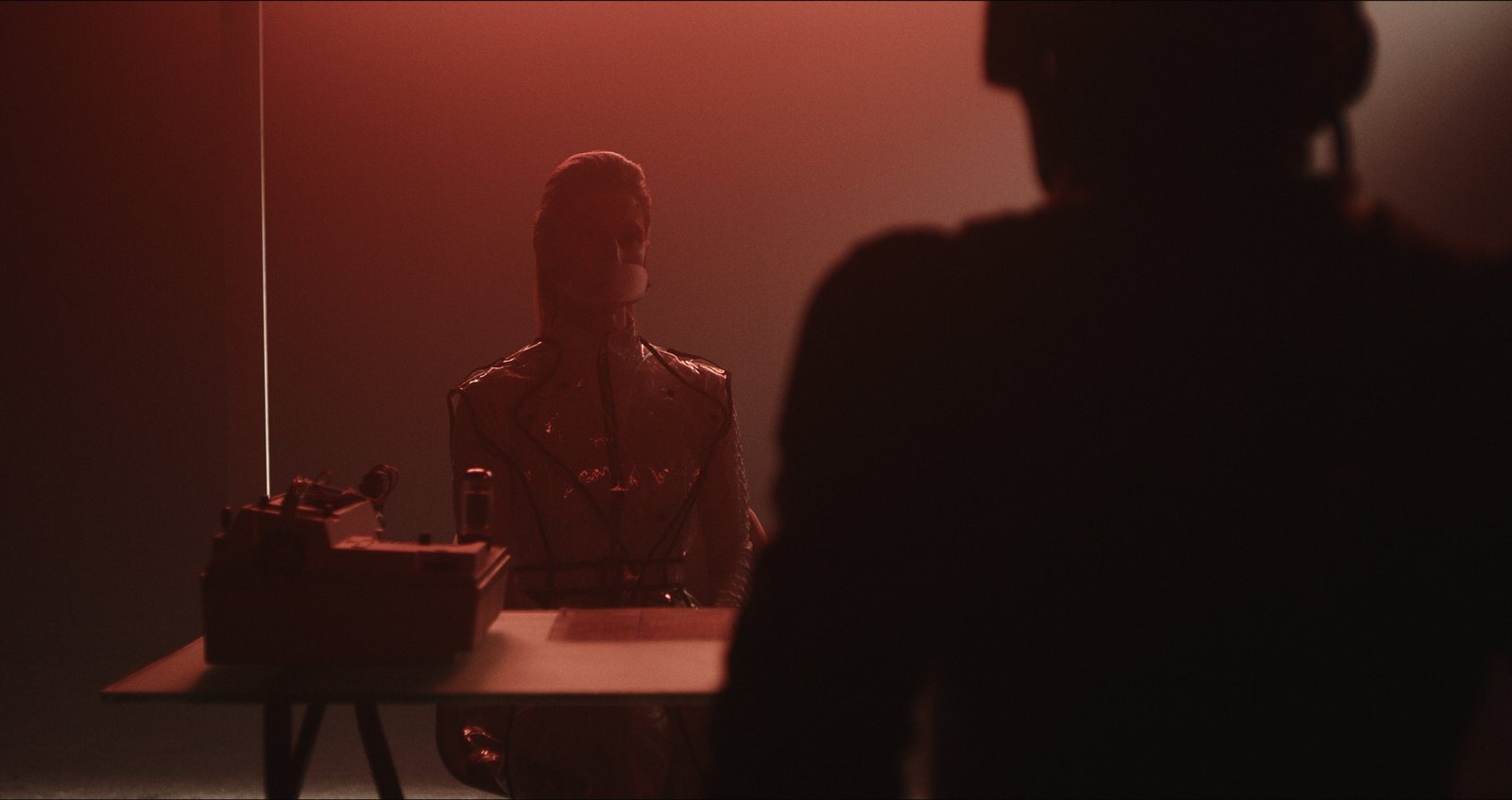 Still from Hello, World: a figure in a glossy suit sits at a table beside an old machine, washed in red light, with a silhouetted figure in the foreground.