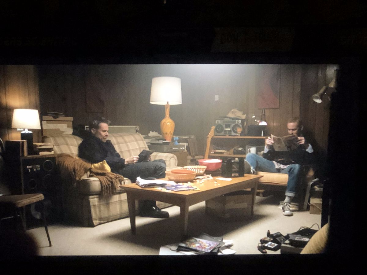 A monitor on set showing the basement as built: wood paneling, a plaid couch and lamps, two men reading comics.