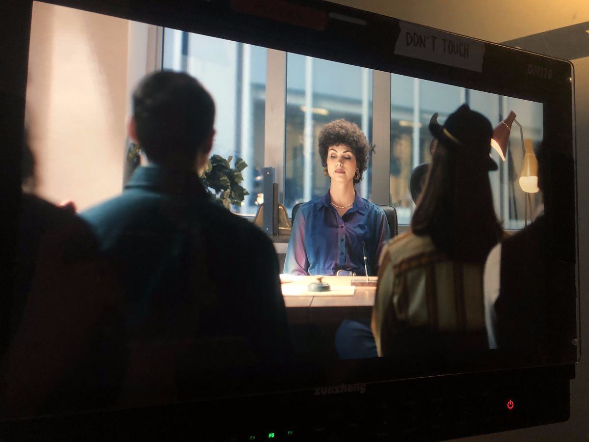 A monitor on set showing a period office in New York: a woman at a desk faces two people across from her.