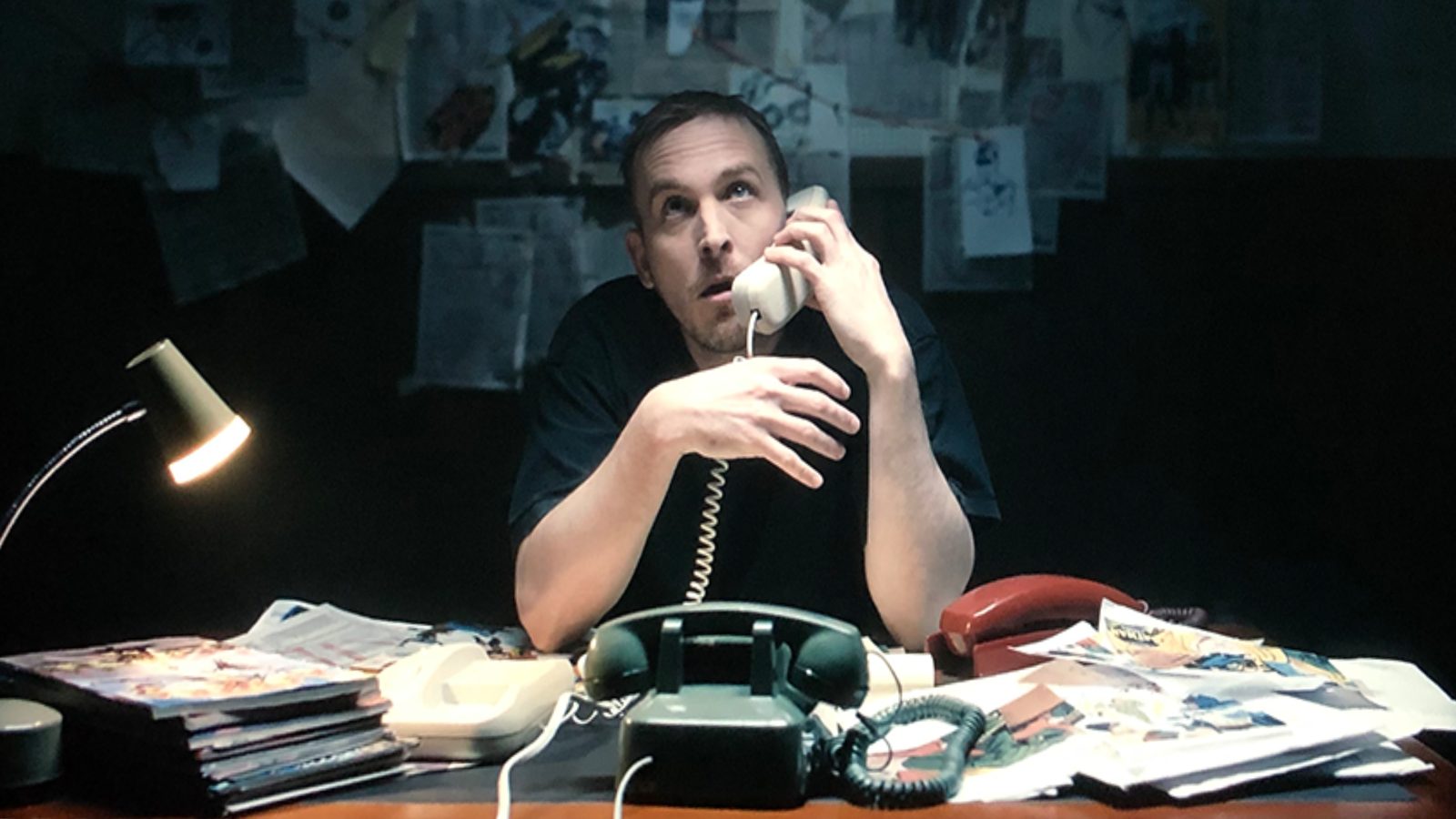 Still from Slugfest. A man sits at a cluttered desk on a corded phone, a wall of pinned papers behind him.