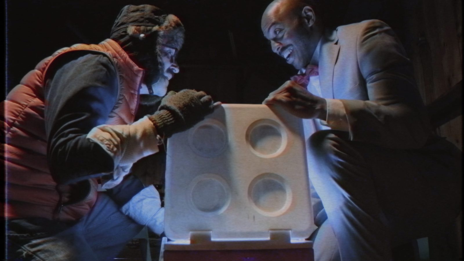 Still from TableTalk. Two people in winter coats and gloves open a white case at night, lit in warm red.